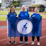 Group picture of 3 people wearing blue active wear, holding a Western Isles Games Association flag.